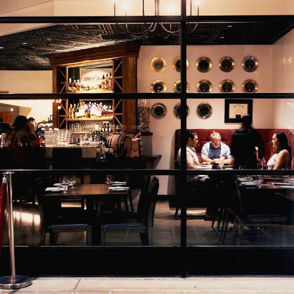 Restaurant interior viewed through glass, featuring a well-stocked bar, dining tables, wall-mounted wine displays, and seated guests.