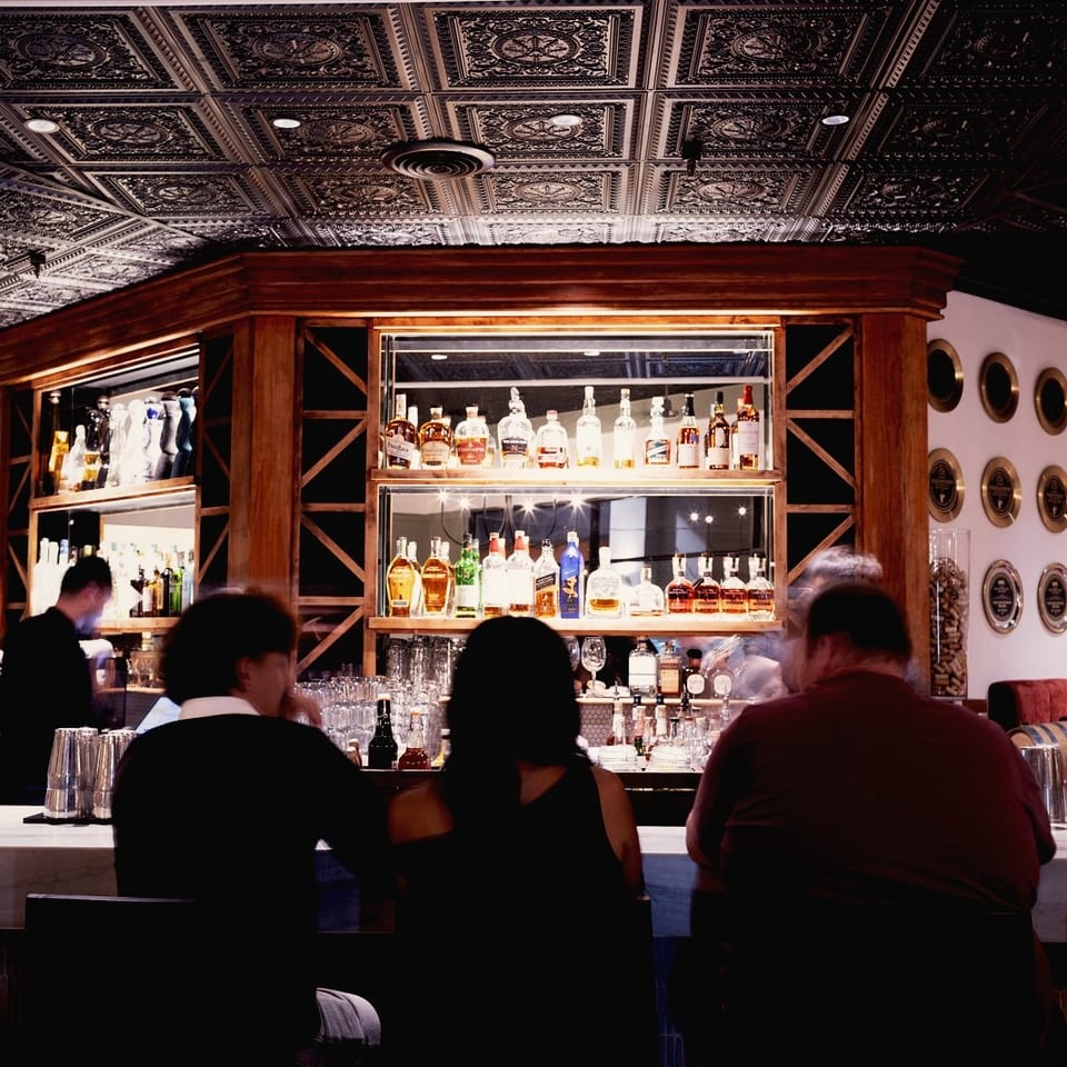 Diners sit at a restaurant bar beneath an ornate ceiling, with illuminated liquor shelves, bartenders, and decorative wall plates.