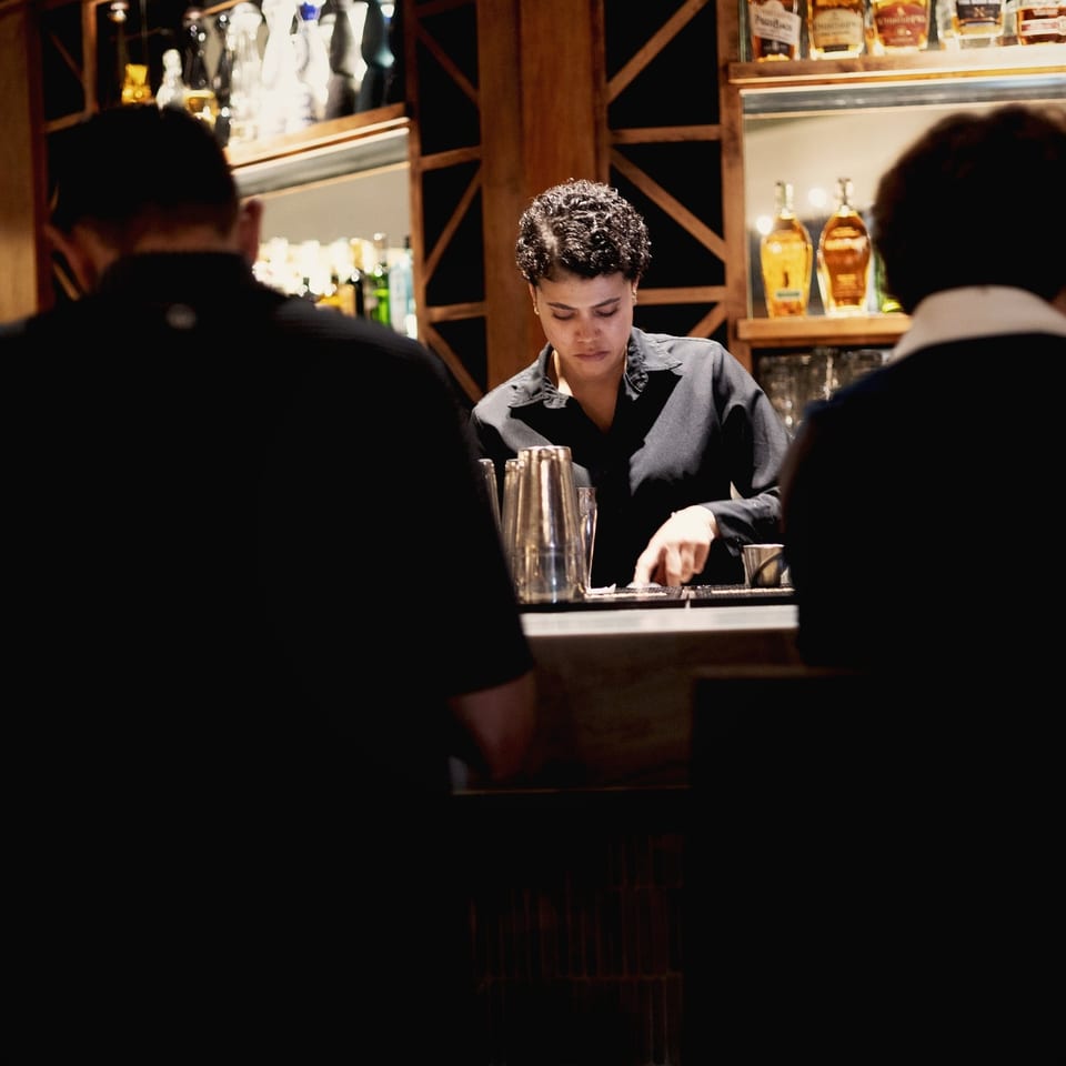 Bartender prepares drinks behind a warmly lit bar stocked with liquor bottles, while two patrons sit in the foreground.