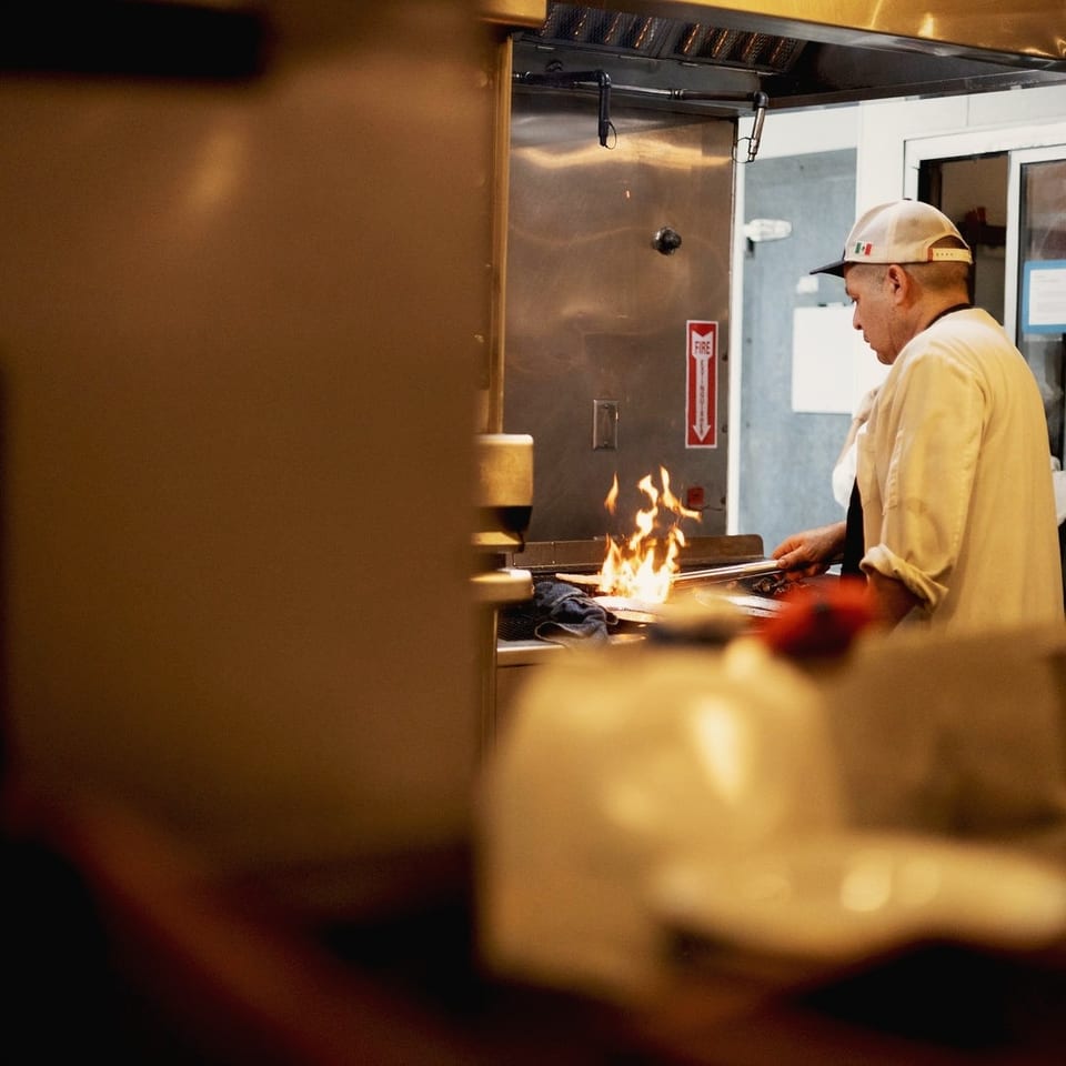 Chef in a white uniform cooks over open flames at a restaurant grill, viewed through the dining area.