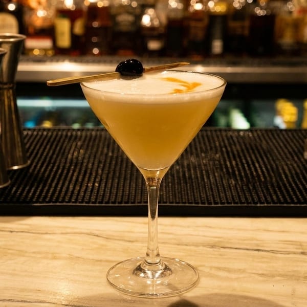 A martini glass with a frothy yellow cocktail, garnished with a cherry, on a bar.
