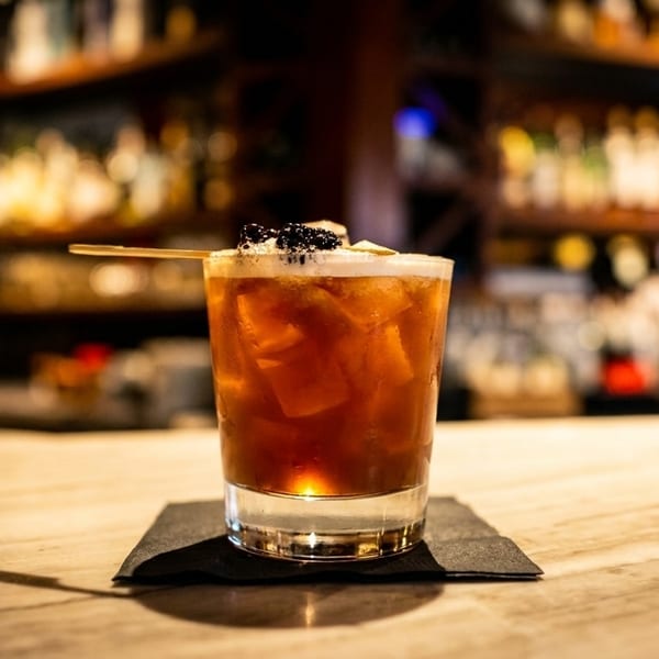 A dark cocktail with ice, topped with foam and blackberries, on a bar with blurred bottles.