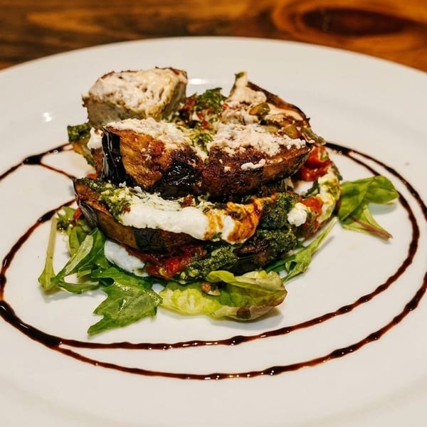 Stacked eggplant parmesan with pesto, cheese, and balsamic glaze on a white plate.