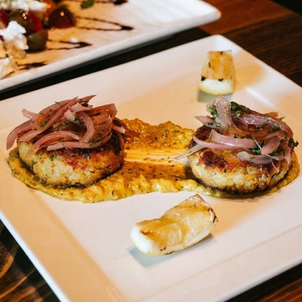 Two crab cakes with pickled red onions and sauce, with another dish in the background.