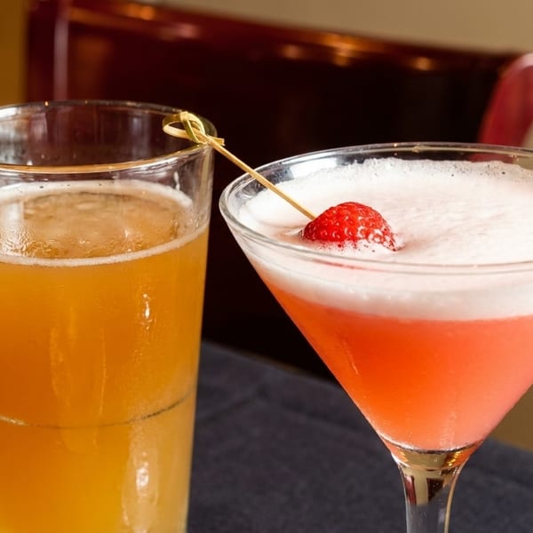 A tall glass of beer next to a pink martini with a strawberry garnish on a dark tablecloth.