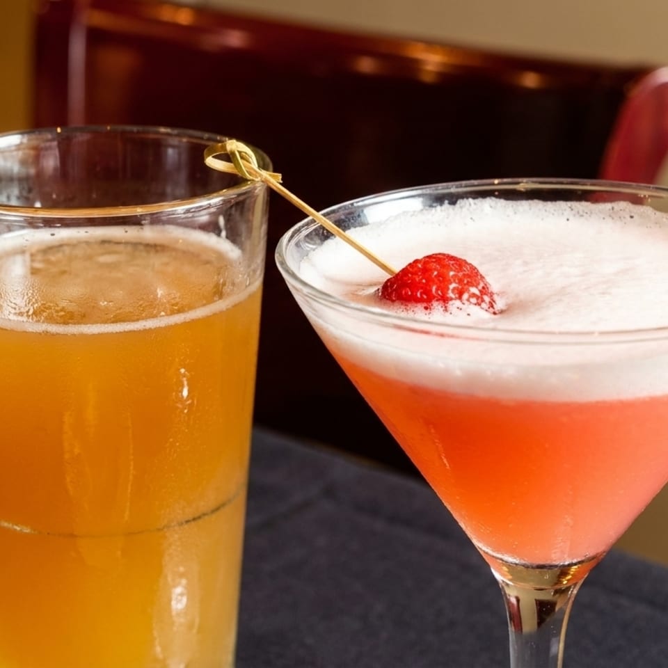 A tall glass of beer next to a pink martini with a strawberry garnish on a dark tablecloth.