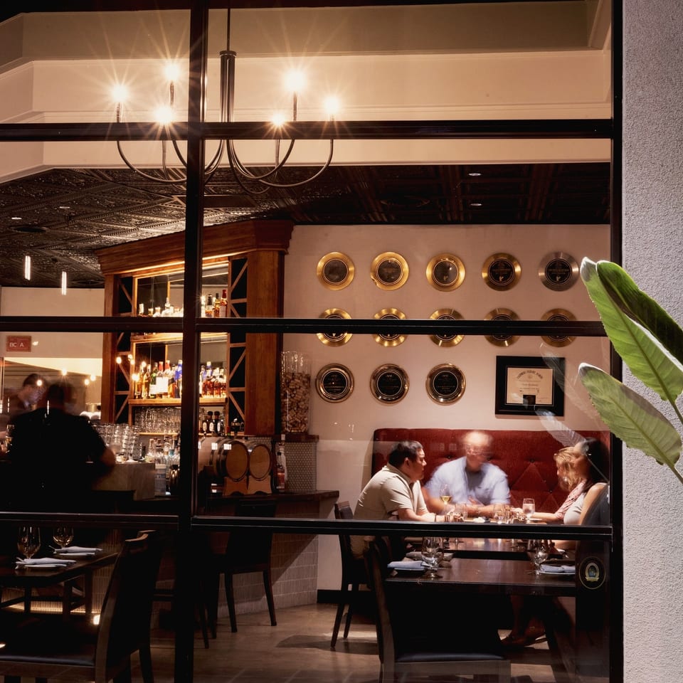 Warmly lit restaurant interior viewed through windows, featuring a bar, diners at tables, chandeliers, and decorative wall plates.